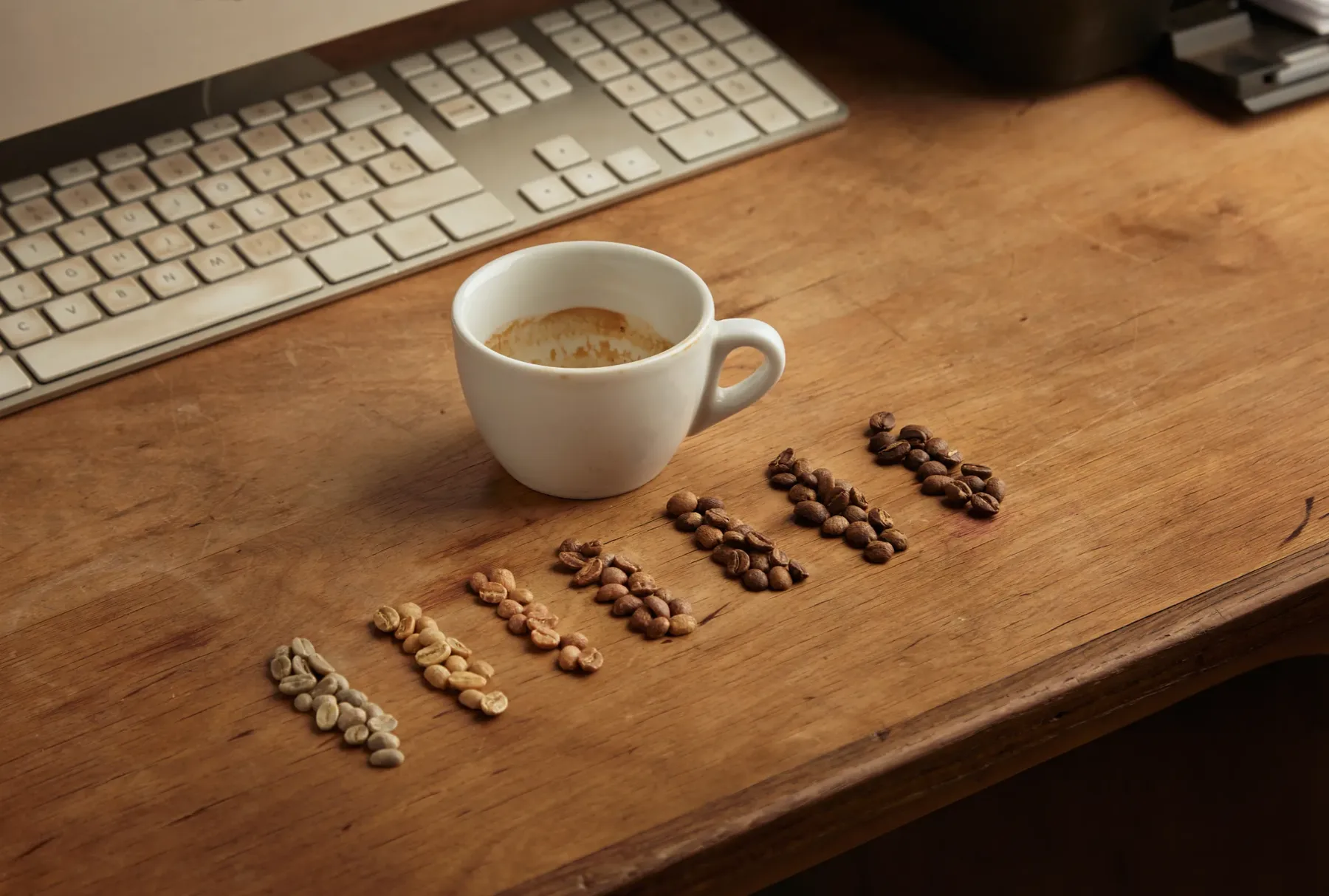 A desk with a keyboard, an empty espresso cup and coffee beans arranged in rising columns, suggesting a daily caffeine routine that builds over time.